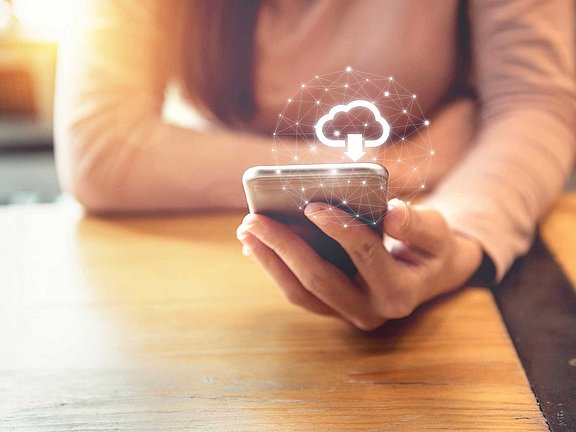 Patientenaufklärung leicht gemacht – Infomaterialien zu Allergiebehandlung & Allergen-Immuntherapie direkt downloaden Eine Person, die ein Smartphone mit einem überlagerten Cloud-Symbol verwendet, das durch Netzwerklinien verbunden ist und Cloud Computing symbolisiert. Die Person sitzt an einem Holztisch und trägt ein helles Langarmshirt.