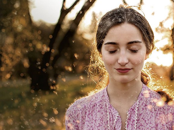 Allergopharma – Ganzheitliche Allergiebehandlung für ein unbeschwertes Leben Eine junge Frau mit geschlossenen Augen und einer rosa Bluse mit Blumenmuster steht draußen in einer ruhigen Umgebung. Hinter ihr geht die Sonne unter und verbreitet einen warmen Schein, während weiche Partikel in der Luft schweben und eine verträumte Atmosphäre schaffen.