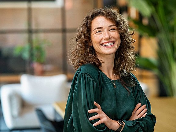 Karriere bei Allergopharma – gemeinsam Zukunft gestalten in der Allergiebehandlung Eine lächelnde Person mit lockigem Haar steht mit verschränkten Armen da und trägt eine dunkelgrüne Bluse. Sie befindet sich im Innenbereich mit einem unscharfen Hintergrund mit Pflanzen und Möbeln.