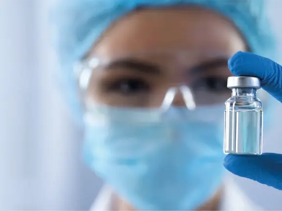 Produktportfolio von Allergopharma. Ein Mitarbeiter im Gesundheitswesen mit blauer Kappe, Gesichtsmaske und Schutzbrille hält ein kleines Fläschchen mit einer klaren Flüssigkeit hoch und konzentriert sich auf das Fläschchen. Der Hintergrund ist verschwommen und hebt das Fläschchen und die behandschuhte Hand hervor.