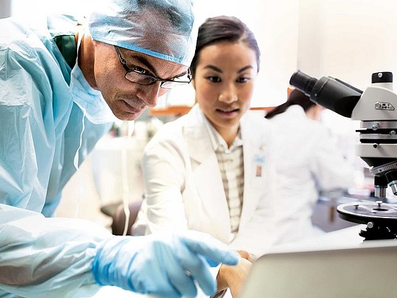 Forschung trifft Praxis – innovative Allergiediagnose für Fachkreise Zwei Wissenschaftler in einem Labor, einer davon in OP-Kittel und Handschuhen, zeigen auf einen Laptop-Bildschirm. Beide wirken konzentriert. Rechts ist ein Mikroskop zu sehen. Der Laborhintergrund ist hell und verschwommen, was auf eine professionelle Forschungsumgebung schließen lässt.